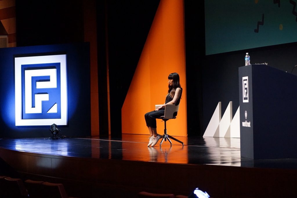 woman sits on a stage in front of lights and logos for a conference